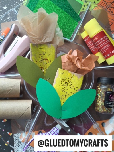 A flat lay of craft supplies, including toilet paper roll corn cob pieces, green and brown paper leaves, glitter, paint, glue, scissors, and a stapler, with a partially assembled cactus craft in the center. @GLUEDTOMYCRAFTS is displayed at the bottom.