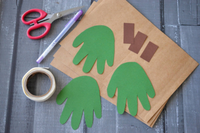 Green hand-shaped cutouts, brown paper, brown rectangles, scissors, a purple pen, and tape are arranged on a gray wooden surface—perfect supplies for creating a festive handprint Christmas tree or making unique handprint Christmas cards for your next craft project.