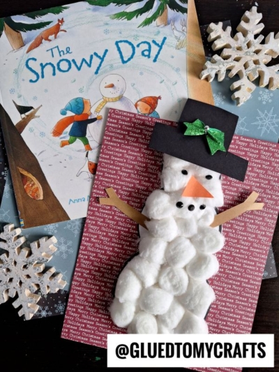A cotton ball snowman craft on red paper with a black hat, carrot nose, and green bow sits next to the book The Snowy Day and two white snowflake decorations, making a charming display of cotton ball snowmen perfect for wintertime fun.
