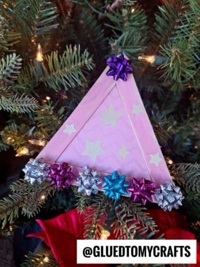 A pink, triangular craft stick party hat ornament—decorated with pastel stars and colorful gift bows—hangs on a lit Christmas tree. @GLUEDTOMYCRAFTS is written at the bottom.