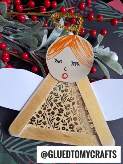 A handmade craft stick angel ornament, also known as a popsicle stick angel, features a paper face with orange hair, gold halo, white paper wings, and a floral dress, beautifully displayed on festive greenery and red berries.