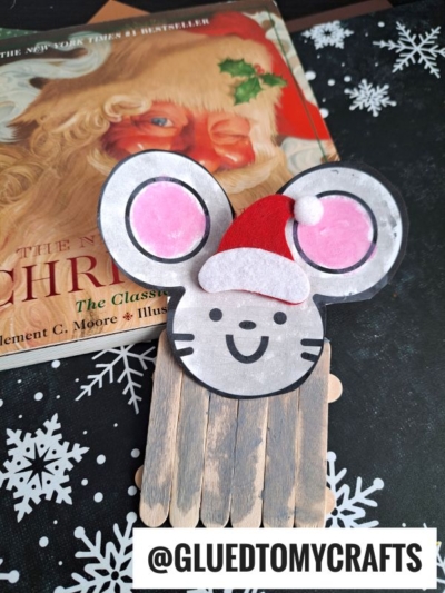 A popsicle stick mouse wearing a Santa hat, crafted from paper and sticks, sits on a snowflake-patterned surface beside a book featuring Santa's face. The text @GLUEDTOMYCRAFTS appears at the bottom.