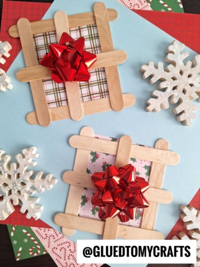 Two popsicle stick present crafts shaped like picture frames with plaid and holly paper centers, each topped with a shiny red bow, are surrounded by decorative snowflakes and layered festive papers. @GLUEDTOMYCRAFTS is at the bottom.