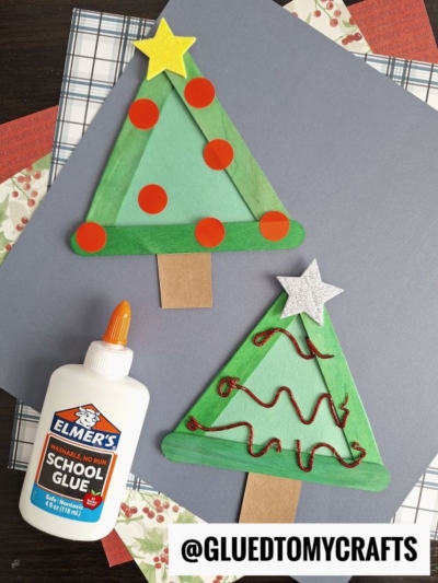 Two popsicle stick Christmas trees, crafted from green sticks with festive decorations, brown paper trunks, and star toppers, sit on blue paper beside a bottle of Elmer’s school glue. Patterned papers decorate the background.