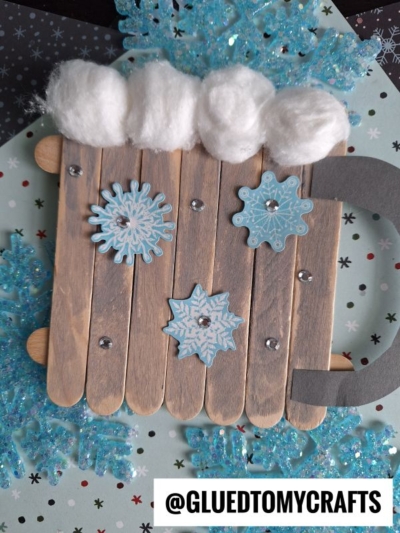 A hot chocolate mug made from wooden craft sticks is decorated with blue snowflakes, white cotton “snow,” and rhinestones, set on sparkling blue snow with the handle on the right—@GLUEDTOMYCRAFTS is written at the bottom.