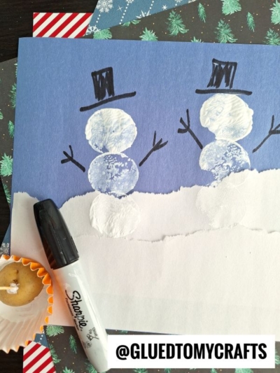 A children’s snowman craft using white paint fingerprints—like potato stamped snowmen—on blue paper, decorated with hand-drawn black hats, arms, and faces. A black marker and a used cupcake liner with paint are nearby. @GLUEDTOMYCRAFTS appears at the bottom.