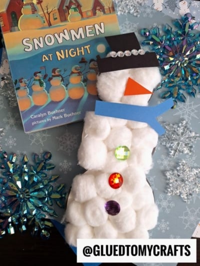 A children’s book titled Snowmen at Night sits beside a cotton ball snowman craft made of cotton balls, colored gems, and paper on a blue snowflake-patterned background. The handle @GLUEDTOMYCRAFTS appears at the bottom.