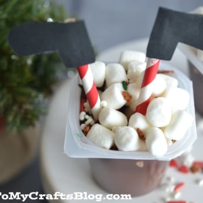 A Santa Stuck In The Chimney Pudding cup is topped with mini marshmallows, a red and white striped straw, and black paper elf legs for a whimsical look. Holiday sprinkles are scattered around to complete the festive treat.