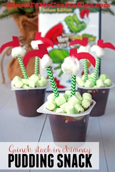 Chocolate pudding cups topped with green mini marshmallows, a green striped straw, and a red paper Grinch leg decoration create a whimsical treat inspired by the Grinch stuck in a chimney—perfect for your Santa Stuck In The Chimney Pudding table. Background includes a Grinch-themed book.