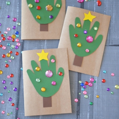Three handmade handprint Christmas cards feature green handprint Christmas trees, decorated with colorful gem stickers and a yellow star. The cards sit on a wooden surface scattered with loose gems. Text reads: Handprint Christmas Tree Cards.