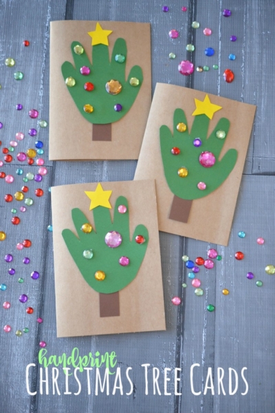 Three handmade handprint Christmas cards feature green handprint Christmas trees, decorated with colorful gem stickers and a yellow star. The cards sit on a wooden surface scattered with loose gems. Text reads: Handprint Christmas Tree Cards.