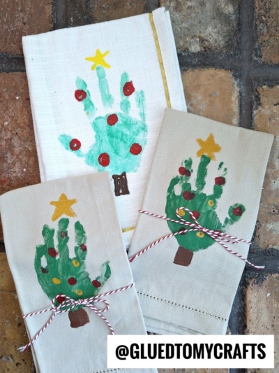 Three hand towels decorated with green handprint Christmas trees, red and yellow ornaments, yellow stars on top—perfect as festive handprint tree napkins. Each is tied with red and white string. Brown tile background. @GLUEDTOMYCRAFTS is displayed at the bottom.