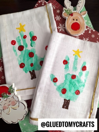 Two white handprint Christmas tree napkins feature green handprints adorned with yellow stars and red ornaments. Nearby, a reindeer and Santa paper decoration add festive charm. The text @GLUEDTOMYCRAFTS is at the bottom.