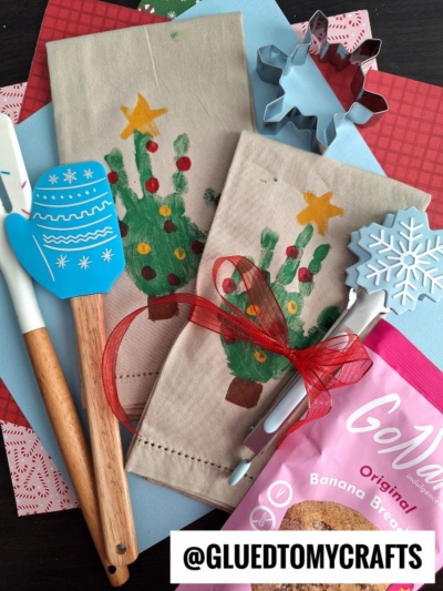 A flat lay of Christmas-themed kitchen items, including spatulas, handprint Christmas tree napkins, cookie cutters, a red ribbon, patterned papers, and a bag of banana bread mix with an @GLUETOMYCRAFTS label.