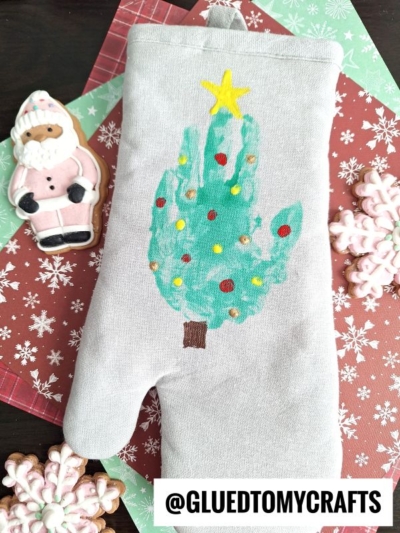 A gray oven mitt with a painted handprint tree decorated with dots and topped with a yellow star rests on festive red and green paper, surrounded by a Santa cookie and pink snowflake cookies. The handle @GLUETOMYCRAFTS is visible.