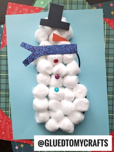 A cotton ball snowman craft features a blue glittery scarf, black hat, orange nose, and colorful buttons, arranged on layered patterned paper. @GLUEDTOMYCRAFTS is written at the bottom. Perfect for cotton ball snowmen fans!.