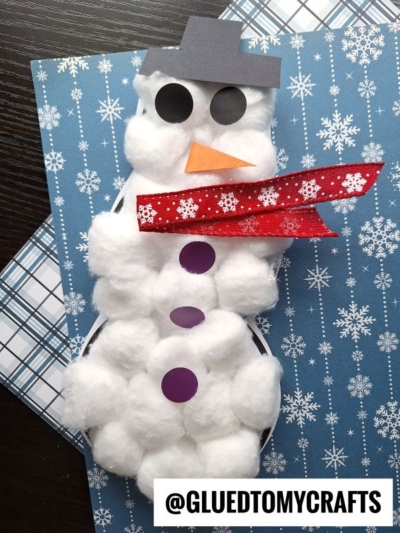 A cotton ball snowman craft featuring paper circles for eyes and buttons, a paper hat and nose, plus a red patterned scarf, set on a blue snowflake background. Text reads @GLUEDTOMYCRAFTS. Perfect idea for cotton ball snowmen fun!.