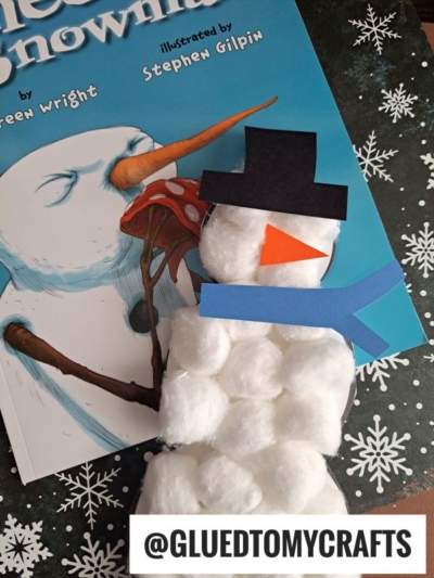 A charming cotton ball snowman with a paper hat, scarf, and nose lies atop a Snowman book. This adorable craft, tagged @GLUEDTOMYCRAFTS, is the perfect inspiration for those wanting to create their own cotton ball snowmen. The scene is set against a festive snowflake-patterned surface.