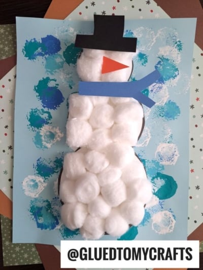 A cotton ball snowman craft made with white cotton balls on blue paper, featuring a black paper hat, blue scarf, and red nose, surrounded by painted blue and white circles. @GLUEDTOMYCRAFTS is at the bottom.