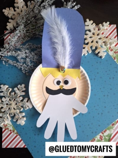 A handmade King Winter craft featuring a paper plate, blue paper hat, white feather, googly eyes, black mustache, yellow crown, and a white handprint beard—perfect inspiration for a festive nutcracker kid craft with snowy blue paper accents.