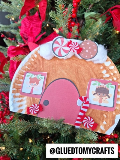 A handmade Paper Plate Gingerbread House craft with candy decorations and two paper characters in the windows is displayed on a Christmas tree with red berries. The Instagram handle @GLUEDTOMYCRAFTS is at the bottom.