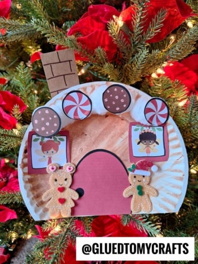 A Paper Plate Gingerbread House craft featuring candy cutouts, windows with cartoon faces, and two gingerbread figures creates a whimsical gingerbread fairy house scene in front of a Christmas tree with red poinsettias. @GLUEDTOMYCRAFTS at the bottom.
