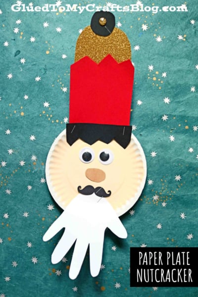 A handmade paper plate nutcracker kid craft featuring a red and gold paper hat, googly eyes, black mustache, and a white handprint beard, displayed on a green starry background. Text reads PAPER PLATE NUTCRACKER.