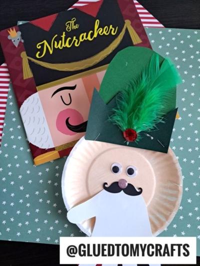 A paper plate Christmas nutcracker kid craft, featuring a green paper hat with feather and red gem, googly eyes, and mustache, sits on star-patterned paper beside a Nutcracker book. @GLUEDTOMYCRAFTS text is at the bottom.