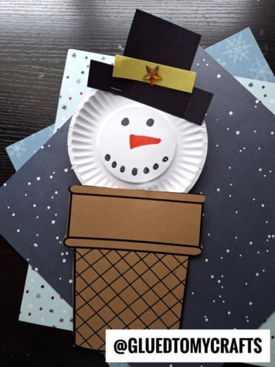 A snowman craft made from a paper plate and colored paper, with the snowman resembling an ice cream cone snowman popping out of a brown paper chimney, set on snowflake-patterned sheets. @GLUEDTOMYCRAFTS is at the bottom.