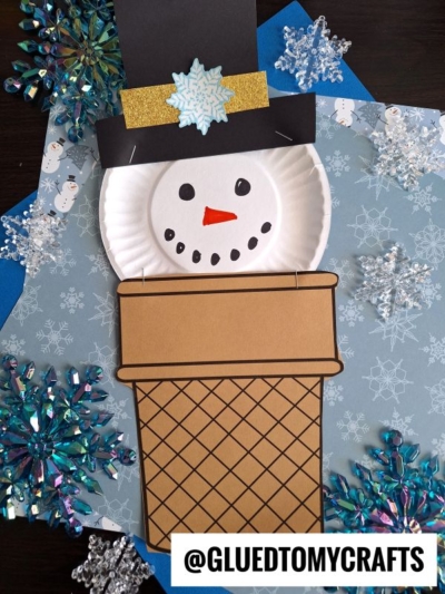A paper craft ice cream cone snowman with a black hat and gold band, made from a paper plate and brown chimney, is surrounded by blue paper, snowflake decorations, and a @GLUEDTOMYCRAFTS label at the bottom right.