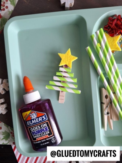 A craft tray holds supplies for a paper straw Christmas tree: green and white striped straws, a clothespin, yellow paper star, purple Elmer’s glue, and a red gift bow. The handle @GLUEDTOMYCRAFTS is also visible.