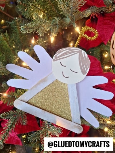 A handmade angel ornament made of white paper handprints, a glittery gold triangle, and a round face with drawn features—just like craft stick angels—hangs on a Christmas tree with red poinsettias. Text reads @GLUEDTOMYCRAFTS.