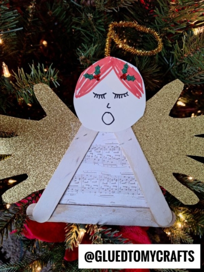 A handmade craft stick angel ornament with gold glitter wings, a drawn face singing, and a music sheet dress hangs on a Christmas tree. The bottom of the image says @GLUEDTOMYCRAFTS.