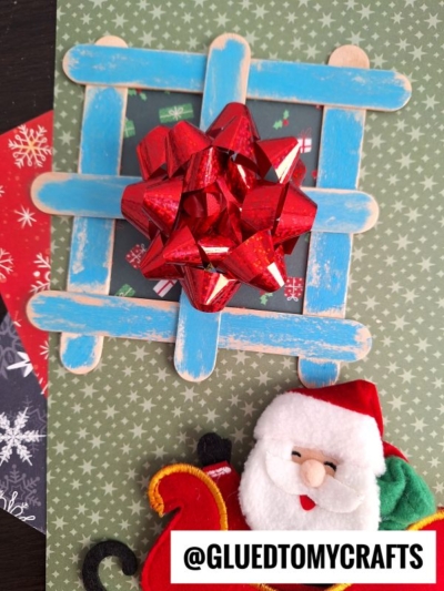 A Christmas-themed craft features a blue popsicle stick present window with a red gift bow in the center, set on green patterned paper. Below is a felt Santa in a sleigh, with “@GLUEDTOMYCRAFTS” printed at the bottom.