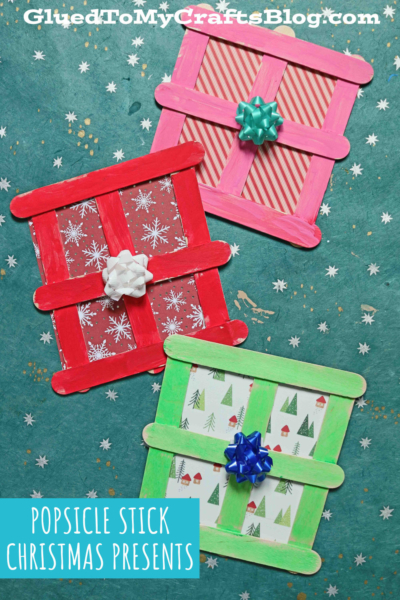 Three square popsicle stick christmas presents made from painted sticks and holiday-patterned paper, each topped with a small gift bow, are displayed on a teal background with white specks. Text reads Popsicle Stick Christmas Presents.