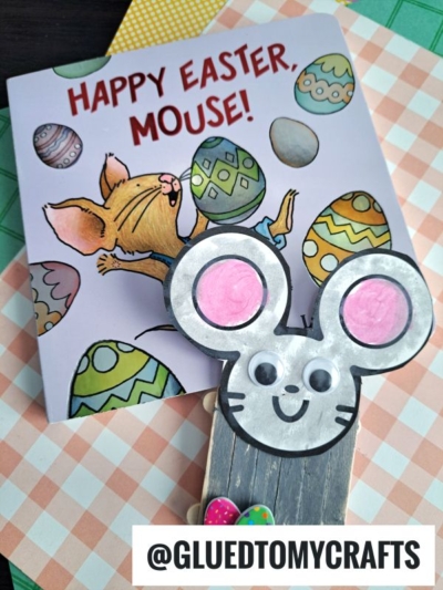 A children’s book titled “Happy Easter, Mouse!” with colorful eggs and a mouse illustration is in the background. In the foreground is a popsicle stick mouse-themed Easter craft made from wood and paper. @GLUEDTOMYCRAFTS is labeled at the bottom.