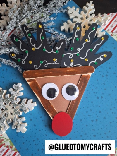 A festive popsicle stick reindeer craft made with a triangle of brown paper, googly eyes, a red nose, and black handprint antlers decorated with ribbon and colorful dots, surrounded by snowflake ornaments. @GLUEDTOMYCRAFTS.
