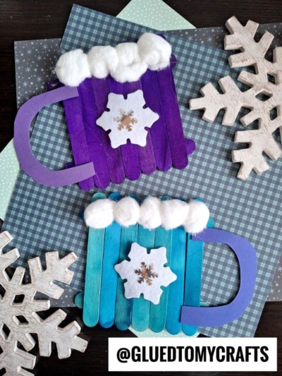 Two handmade popsicle stick hot cocoa mugs, one purple and one blue, are decorated with cotton ball whipped cream and a white felt snowflake. They sit on patterned paper with wooden snowflakes. @gluedtomycrafts is written at the bottom.