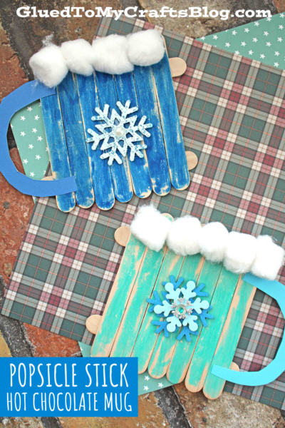 Two popsicle stick hot chocolate mugs, each with a blue handle, cotton ball whipped cream, and snowflake embellishments, rest on a plaid surface. Text reads: POPSICLE STICK HOT CHOCOLATE MUG.