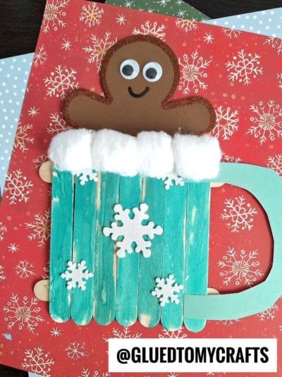 A handmade Popsicle Stick Hot Chocolate Mug features a turquoise mug crafted from popsicle sticks, topped with cotton balls and a brown gingerbread figure with googly eyes. White snowflake decorations and holiday-themed paper complete the festive look.