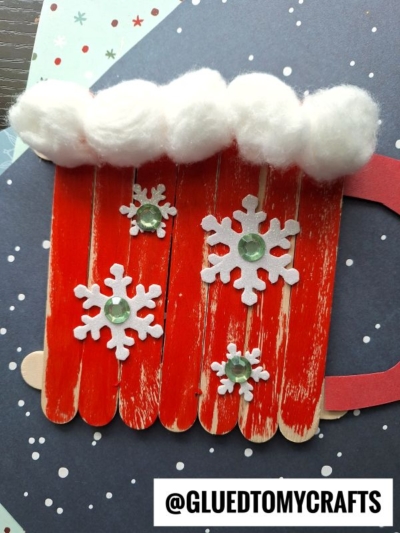 A Popsicle Stick Hot Chocolate Mug craft made from red-painted sticks, decorated with cotton ball foam, snowflake cutouts, and green gems, displayed on a star-patterned background. Text reads @GLUEDTOMYCRAFTS.
