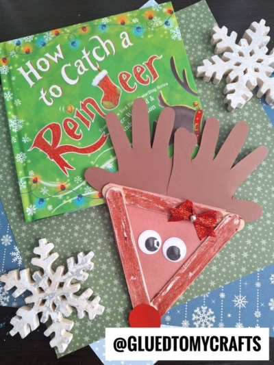 Children’s book “How to Catch a Reindeer” beside a popsicle stick reindeer craft with paper, googly eyes, and a red bow, displayed on a festive green and blue background with snowflake decorations.
