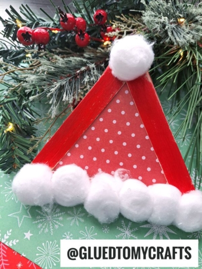 Popsicle Stick Santa Hats handmade from red popsicle sticks, red and white polka-dotted paper, and cotton balls, arranged on green festive paper with pine branches and red berries in the background. Text: @GLUEDTOMYCRAFTS.
