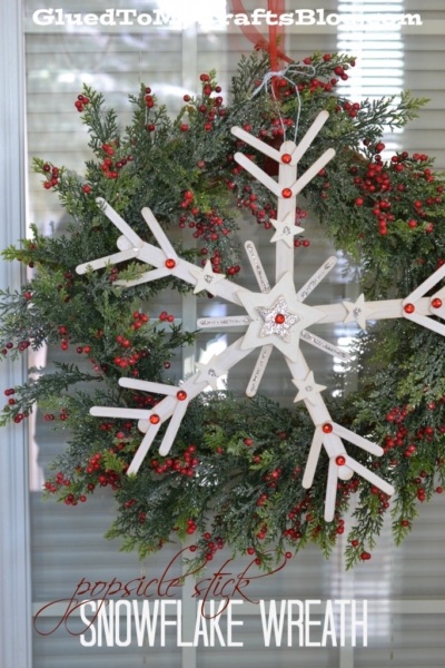 A holiday wreath decorated with green pine branches and red berries displays a handmade popsicle stick snowflake ornament with red gems at its center. Text reads “popsicle stick snowflake wreath.”.