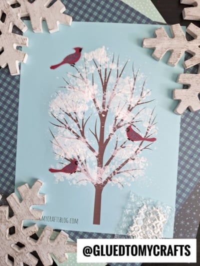 Craft project featuring a printed tree with two red cardinals on light blue paper, accented with bubble wrap snowy tree effects and textured white paint as snow. Surrounded by wooden snowflakes and patterned craft papers. @GLUEDTOMYCRAFTS is labeled at the bottom.