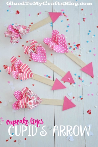 Four Valentine’s Day cupid’s arrows made from wooden sticks, pink paper, and colorful cupcake liners are arranged on a white surface with scattered heart-shaped confetti. Text reads cupcake liner CUPIDS ARROW.