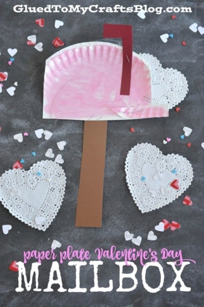 A pink paper plate mailbox craft with a brown post, a red flag, and white lace heart decorations, surrounded by heart-shaped confetti on a gray background. Text reads: Paper Plate Mailbox – the perfect Valentine’s Day project for kids!.