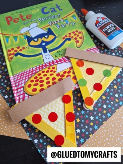 A children’s book titled Pete the Cat and the Perfect Pizza Party sits on a table with a bottle of Elmer’s School Glue and two craft stick pizza slices decorated with colored circles for toppings.
