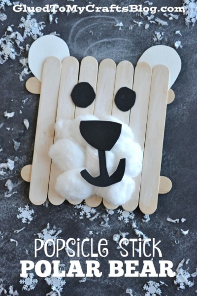 A popsicle stick polar bear face made from craft sticks, cotton balls, and black and white paper, with snowflake decorations surrounding it. Text reads Popsicle Stick Polar Bear.