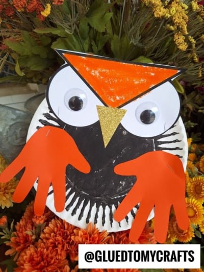 Paper plate owl craft with orange paper wings, large googly eyes, and an orange and gold beak, displayed against colorful artificial flowers. This adorable owl craft for kids is perfect for creative afternoons! @GLUEDTOMYCRAFTS is written at the bottom.